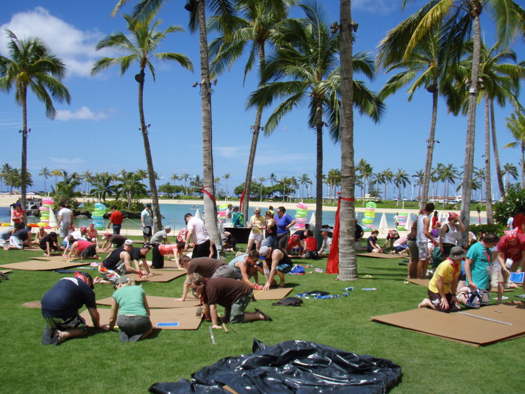Teams constructing cardboard boats on a sunny lawn surrounded by palm trees during the Build-A-Boat team building event.