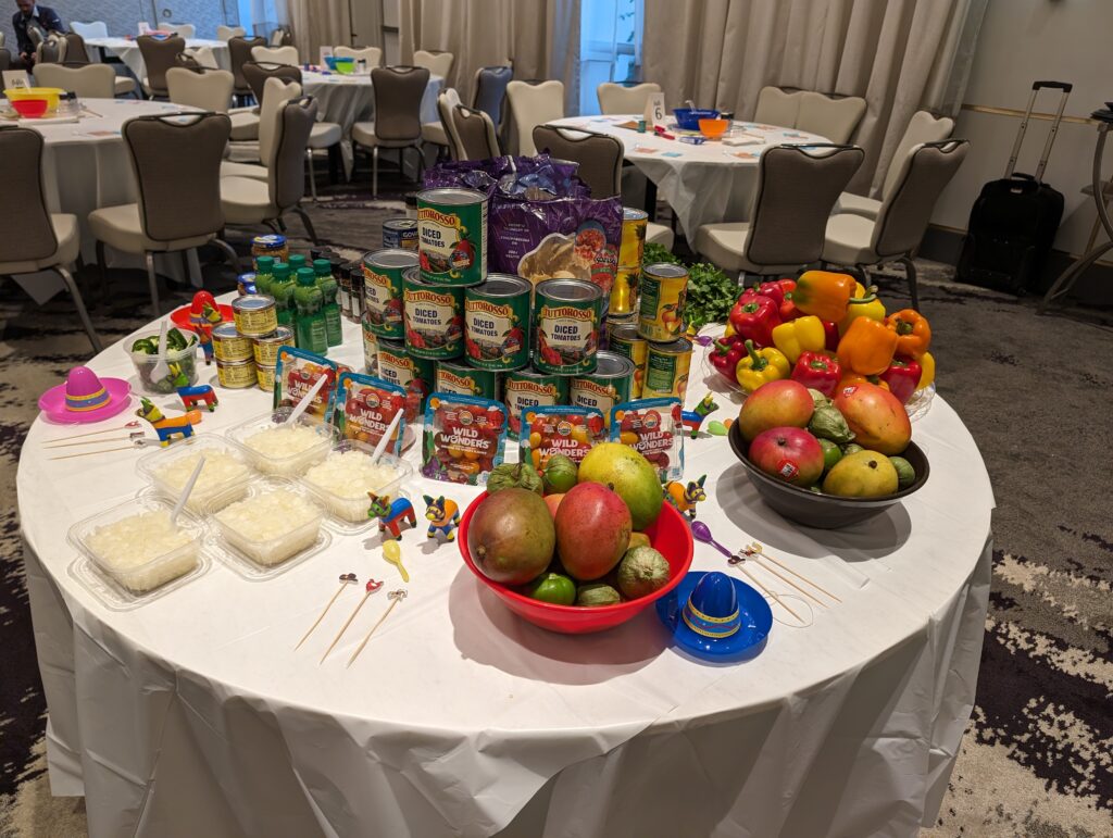 A vibrant setup of fresh fruits, including mangoes and peppers, along with canned ingredients for the Salsa Showdown team building event, featuring colorful decorations.