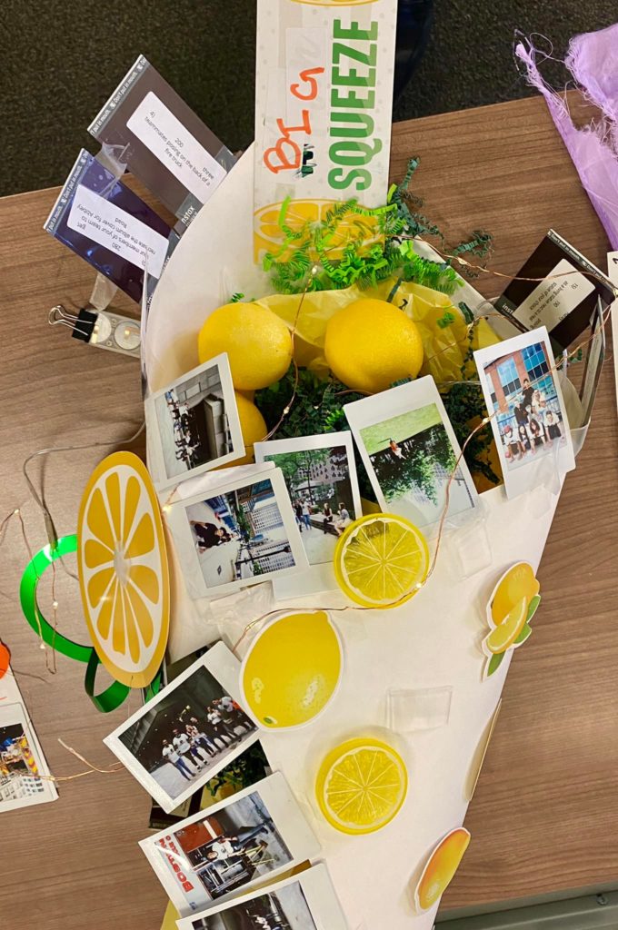 A festive and creative lemon-themed display from a Polaroid scavenger hunt event. The arrangement includes vibrant yellow lemons, greenery, and Polaroid photos scattered across a board, surrounded by lemon cutout decorations and playful text that reads 'Squeeze.' The display captures fun team moments of this team building activity.