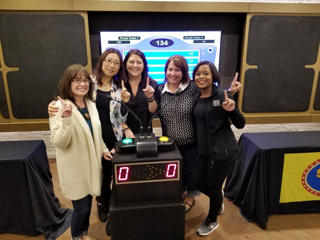 Five participants smiling and holding up their index fingers in front of a game show podium, celebrating their involvement in the Corporate Feud team building activity, with a game board displayed in the background.