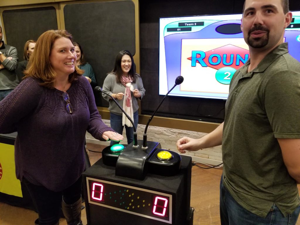 Two participants standing at a game show podium with buzzers, smiling and preparing to answer during the survey game show team building event, with other participants watching in the background.