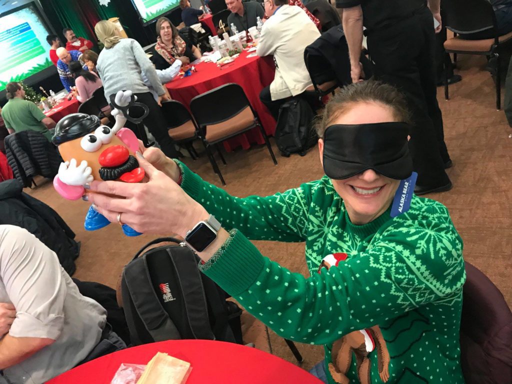 A person wearing a green holiday sweater sits at a table in a large room filled with people. They are holding up a Mr. Potato Head toy while wearing a blindfold, smiling as they participate in an activity. Other attendees in the background are engaged in similar team building holiday-related activities, with festive decorations and red tablecloths creating a cheerful atmosphere.