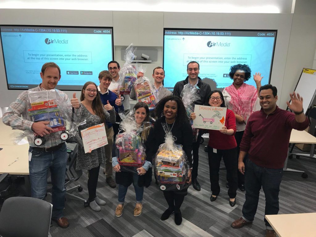 A group of people standing together in a classroom or office setting, smiling and holding gift baskets wrapped in clear plastic. The baskets contain toys, as part of a charitable donation team building event. The group appears to be celebrating their efforts, with some holding signs, and the atmosphere is one of teamwork and achievement.