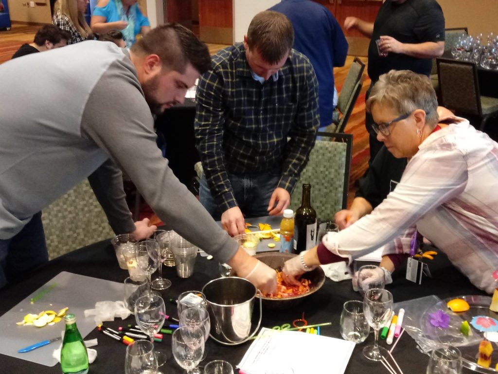 A group of participants actively mixes fruit and ingredients in a large bowl during the 'Winery Team Challenge' event. They are focused on creating their unique sangria blend, using various fruits and drinks on the table. Wine glasses, bottles, and additional supplies are scattered on the table, emphasizing the hands-on nature of the activity. The atmosphere is lively, with other teams visible in the background.