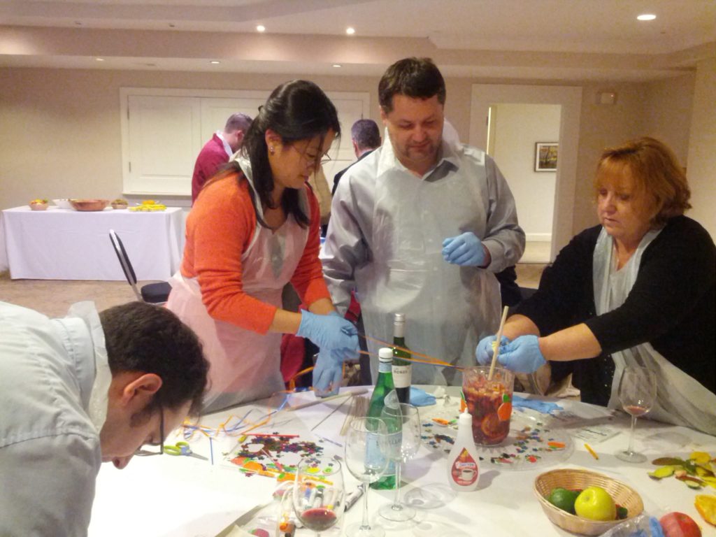 Participants in the 'Winery Team Challenge' work together at a table, wearing aprons and blue gloves as they prepare their signature sangria. The table is covered with colorful decorations, fresh fruit, and bottles, with a pitcher of sangria in the center. The team members appear focused and collaborative, contributing to various aspects of the sangria-making process, from mixing ingredients to adding finishing touches. This image captures the hands-on, creative spirit of the team building event.