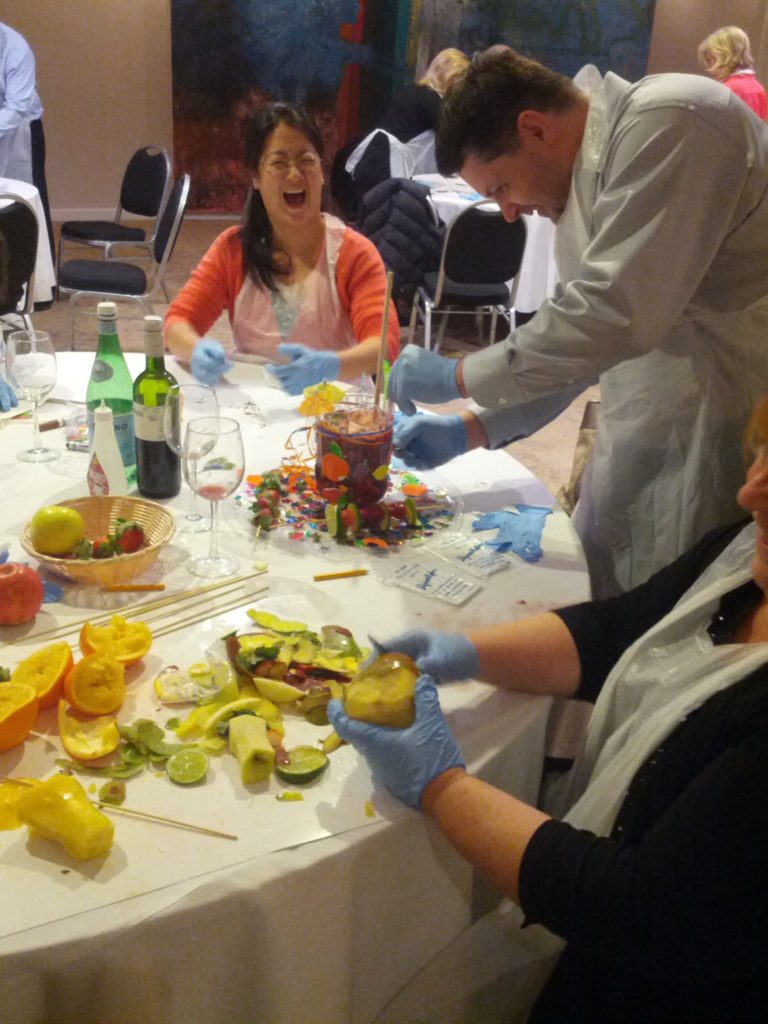 Three participants enthusiastically engage in the 'Winery Team Challenge' team building event. The group is gathered around a table, laughing and preparing their unique sangria blend. One person in the foreground is holding a piece of fruit and wearing blue gloves, while another participant mixes ingredients into a large, decorated sangria pitcher. The table is filled with sliced fruits, wine bottles, and colorful decorations, adding to the fun and creative atmosphere of the event.