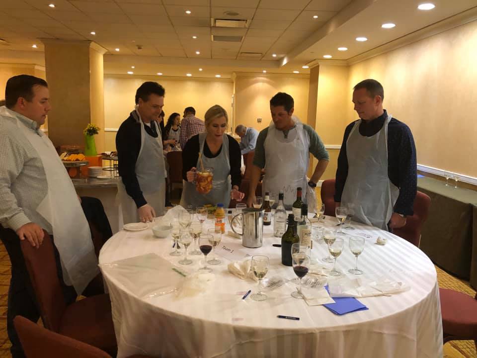 A group of five participants stand around a round table covered with a white tablecloth during the 'Winery Team Challenge.' They are wearing aprons and discussing their next steps as one participant holds a pitcher filled with a colorful sangria blend. The table is scattered with empty wine glasses, bottles of wine, and various ingredients like juices and spices, creating an atmosphere of teamwork and creativity as they finalize their unique sangria creation.