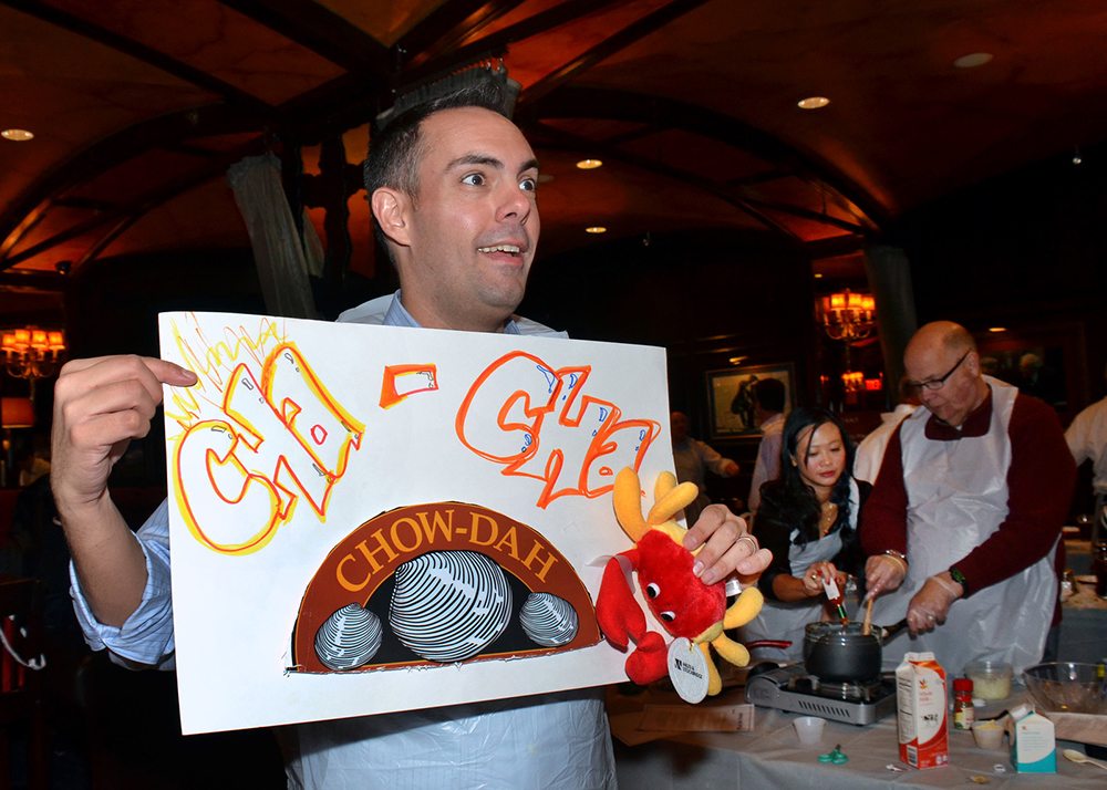 Participant enthusiastically holding a ‘Cha-Cha Chow-dah’ sign during the Wicked Good Chowda Cook-Off team building event.