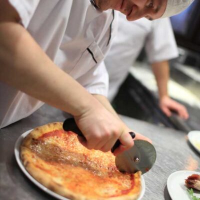 A participant cutting a freshly baked pizza with a pizza cutter during The Ultimate Pizza Challenge team building event.