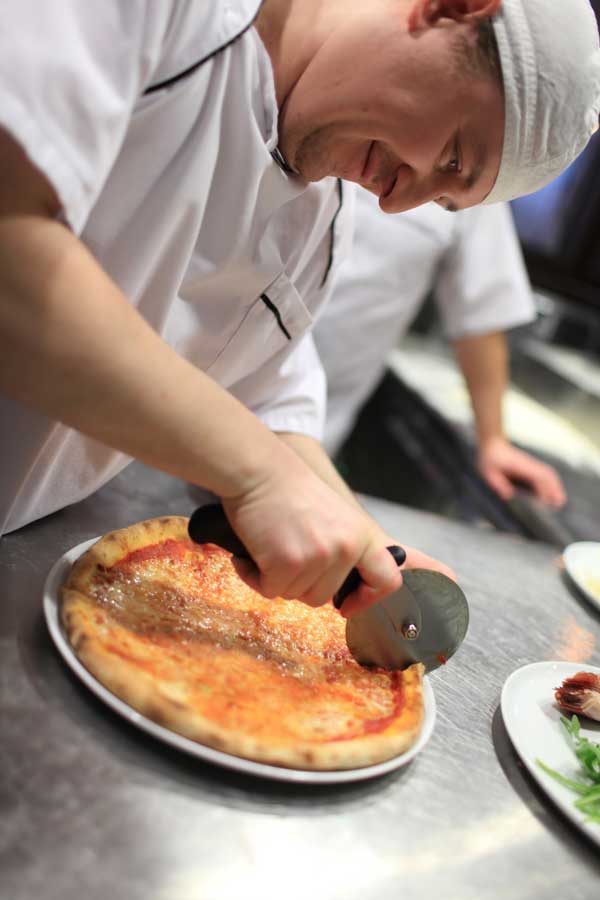 A participant cutting a freshly baked pizza with a pizza cutter during The Ultimate Pizza Challenge team building event.