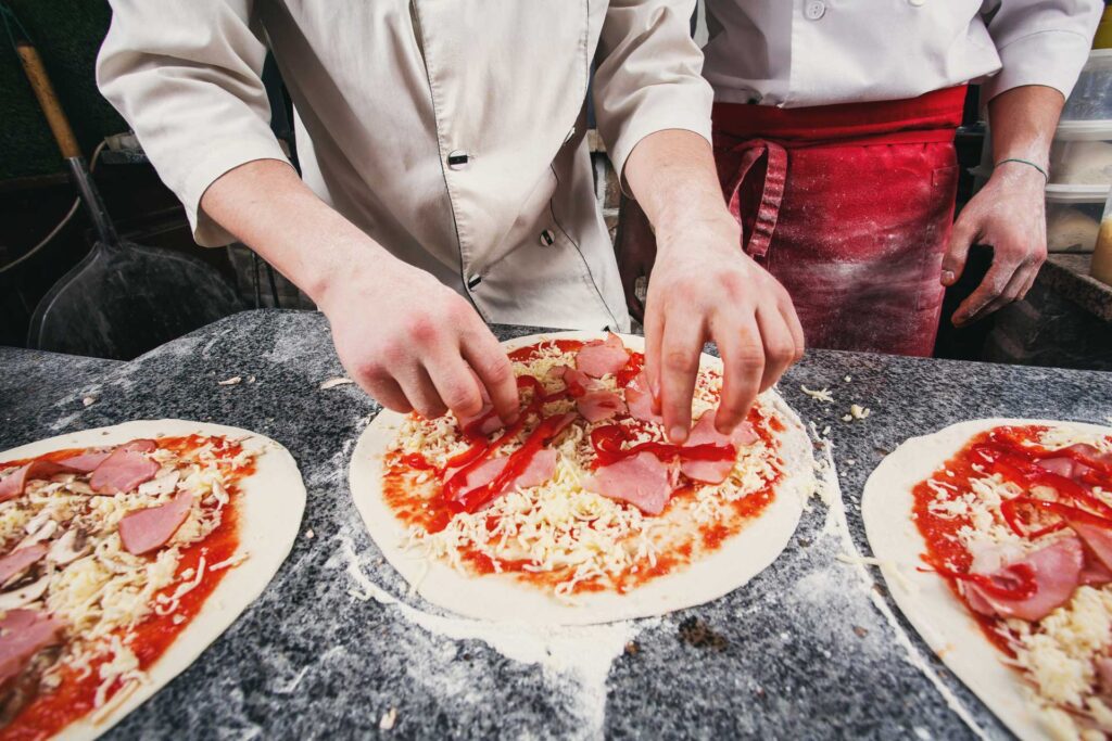 Hands of two participants assembling toppings on pizza dough during The Ultimate Pizza Challenge team building event, focused on culinary teamwork and competition.
