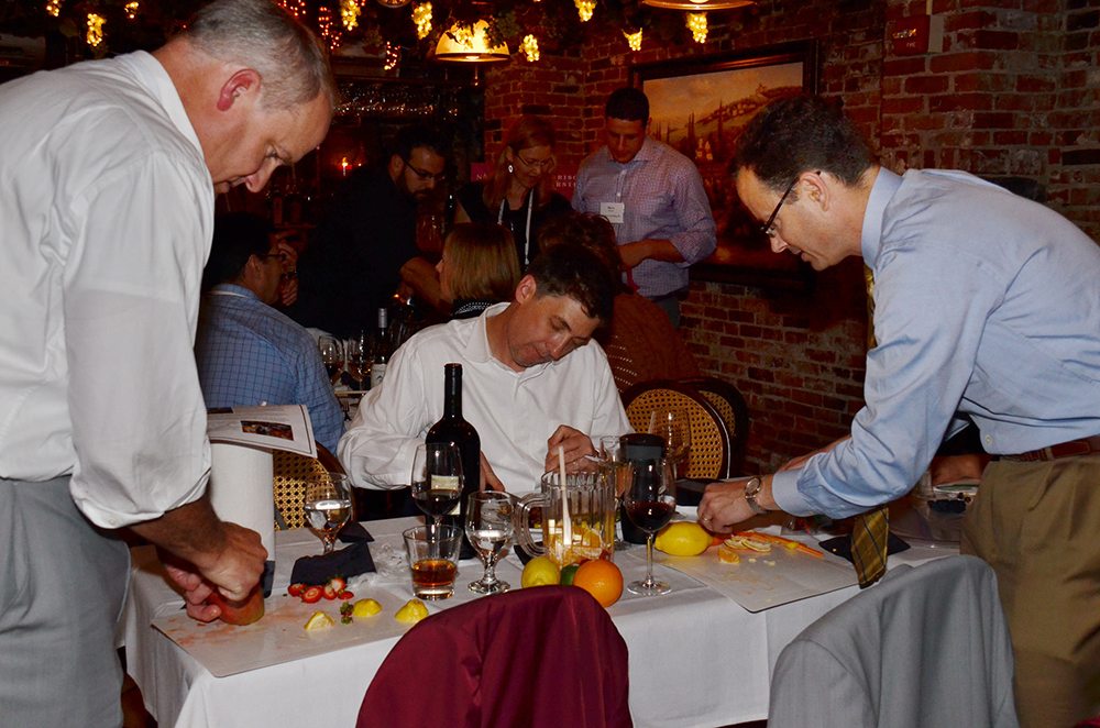Three participants in the 'Winery Team Challenge' event are focused on preparing ingredients for their sangria creation. Two men are cutting various fruits, such as oranges and lemons, at a table with a bottle of wine, glassware, and other supplies. Another participant seated in the middle is reviewing notes. The event is set in a cozy, warmly lit restaurant with brick walls, as other teams can be seen working in the background, creating a lively and collaborative atmosphere.