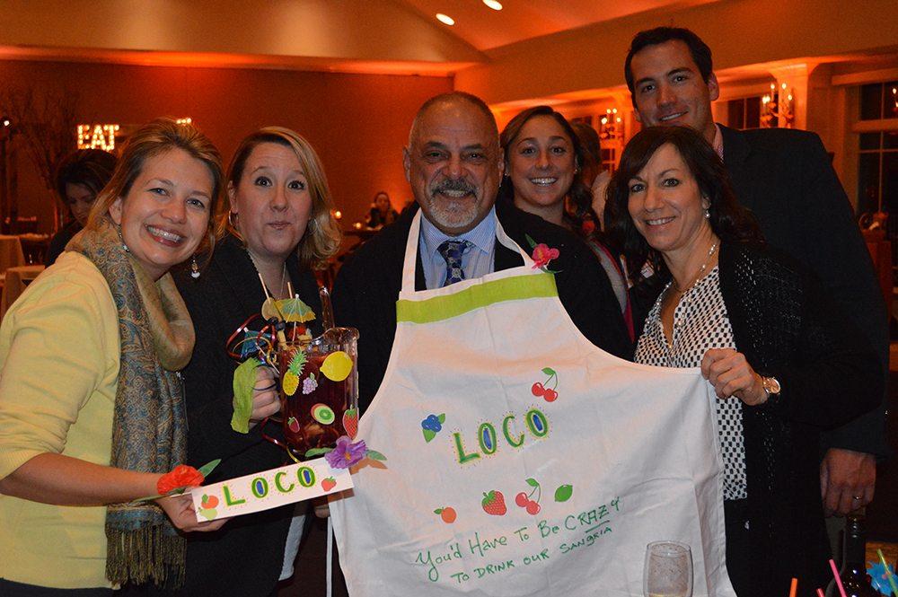 A group of six people smiles and poses for a photo during the 'Winery Team Challenge' event. One person proudly holds a handmade, decorative sign labeled 'LOCO,' while another displays a customized apron with the same name and colorful designs of fruit and flowers. One team member holds a pitcher of sangria garnished with fruit, complete with a fun cocktail umbrella. The atmosphere is warm, with soft lighting in the background, emphasizing the friendly and creative spirit of the team building activity.