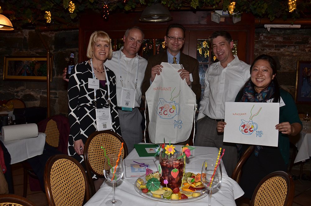A group of five participants in the 'Winery Team Challenge' event proudly display their team's creations. Two of the members hold up custom-designed aprons and logos for their sangria blend. The group stands around a table adorned with a colorful pitcher of sangria surrounded by decorative fruit and festive drink umbrellas. The setting is a cozy, warmly lit venue with rustic brick walls and grapevine decorations hanging from the ceiling, adding to the wine-themed ambiance.