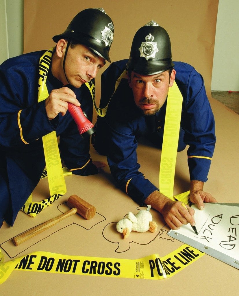 Two actors dressed as police officers humorously investigate a mock crime scene in a playful setup for a Murder Mystery Dinner event. They are kneeling on the ground, surrounded by yellow police line tape, with a chalk outline on the floor resembling the shape of a duck. One officer is holding a red flashlight while the other points at a toy duck with a sign labeled 'Dead Duck.' The scene is lighthearted and comedic, emphasizing the fun, interactive nature of the mystery event.