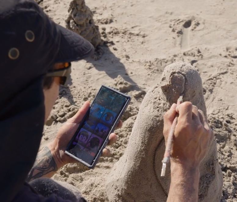 A participant refining their sand sculpture while using a reference image on their phone during a sand sculpting team building event.