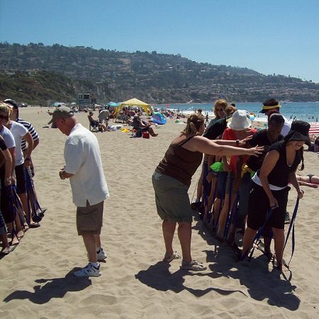 A group of participants collaborating in a sand-skiing relay race during a sand sculpting team building event on the beach.