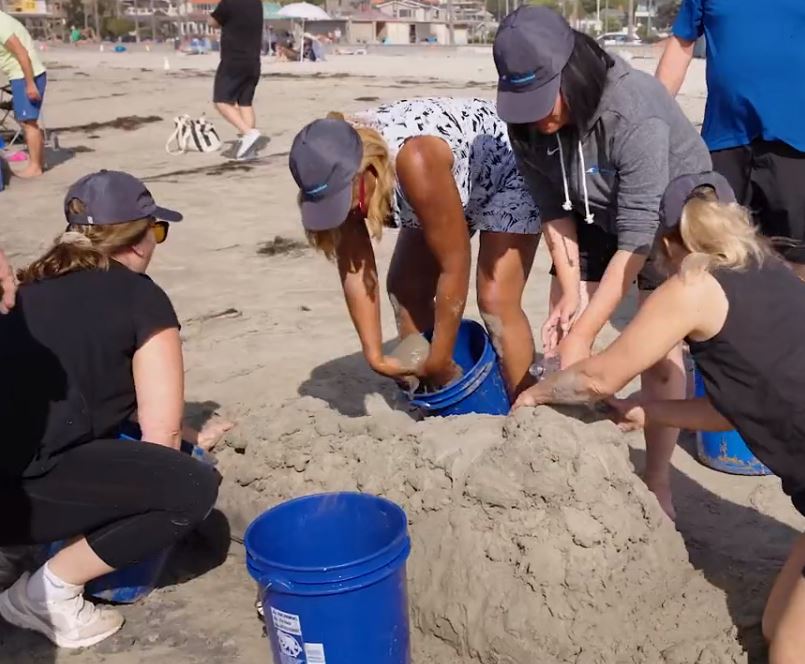 Team collaborating on shaping a sand sculpture using buckets of sand at a sand sculpting team building event on the beach.