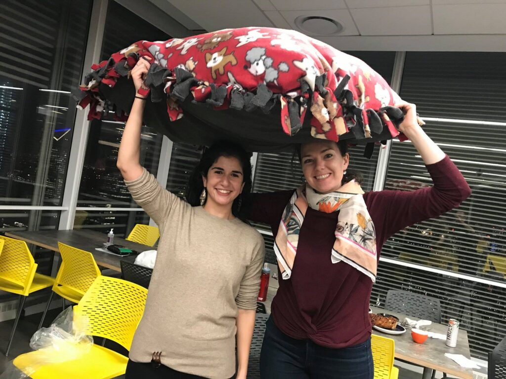 Two smiling participants hold up a large, colorful dog bed they created during the Paws For A Cause team building event, contributing to local animal shelters.