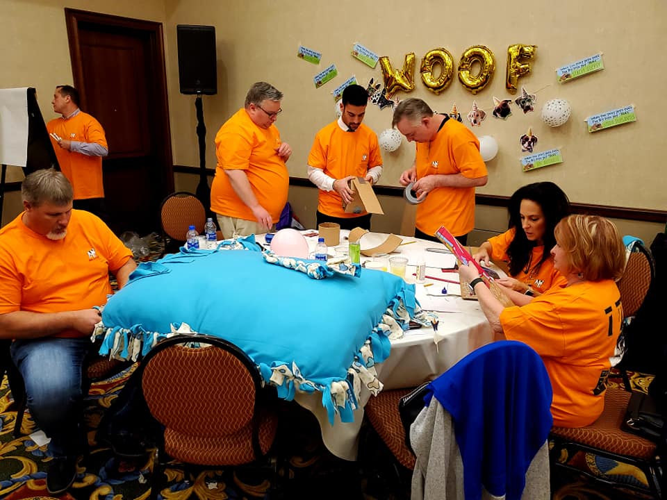 A team dressed in matching orange shirts focuses on crafting a large dog bed and other items during the Paws For A Cause team building event, contributing to local animal shelters.