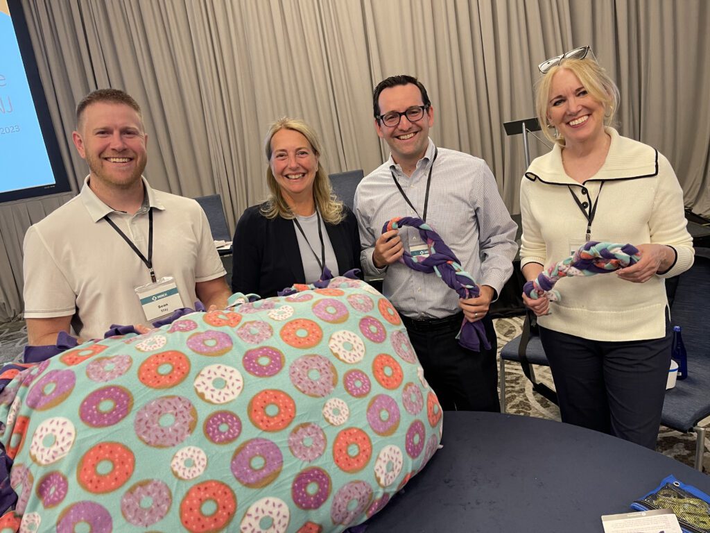 A group of participants from the Paws For A Cause team building event proudly display the dog beds and toys they created, ready to be donated to local animal shelters, with a beautiful waterfront backdrop.