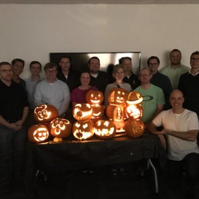 A group of team members proudly display their lit jack-o'-lanterns after a pumpkin carving team building event. The glowing pumpkins feature a variety of creative and fun designs, and the group stands smiling together in a dimly lit room, celebrating their achievements.