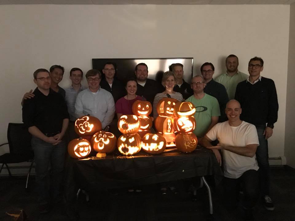 A group of team members proudly display their lit jack-o'-lanterns after a pumpkin carving team building event. The glowing pumpkins feature a variety of creative and fun designs, and the group stands smiling together in a dimly lit room, celebrating their achievements.