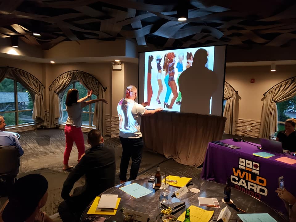 Participants engage in a lively music and dance challenge during the Wild Card Team Building Bingo event, with a vibrant video displayed on the screen.