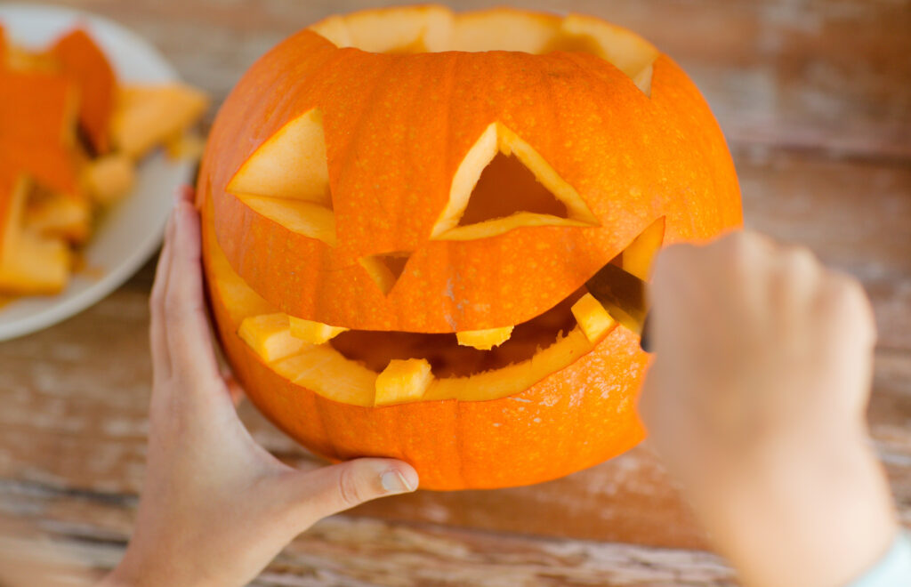 Hands carefully carving a pumpkin, shaping a smiling face with triangular eyes and a wide mouth. Pieces of pumpkin are seen on a plate in the background, showcasing the creative process during a pumpkin carving activity.