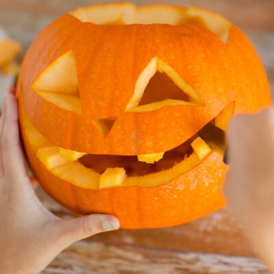 Hands carefully carving a pumpkin, shaping a smiling face with triangular eyes and a wide mouth. Pieces of pumpkin are seen on a plate in the background, showcasing the creative process during a pumpkin carving activity.