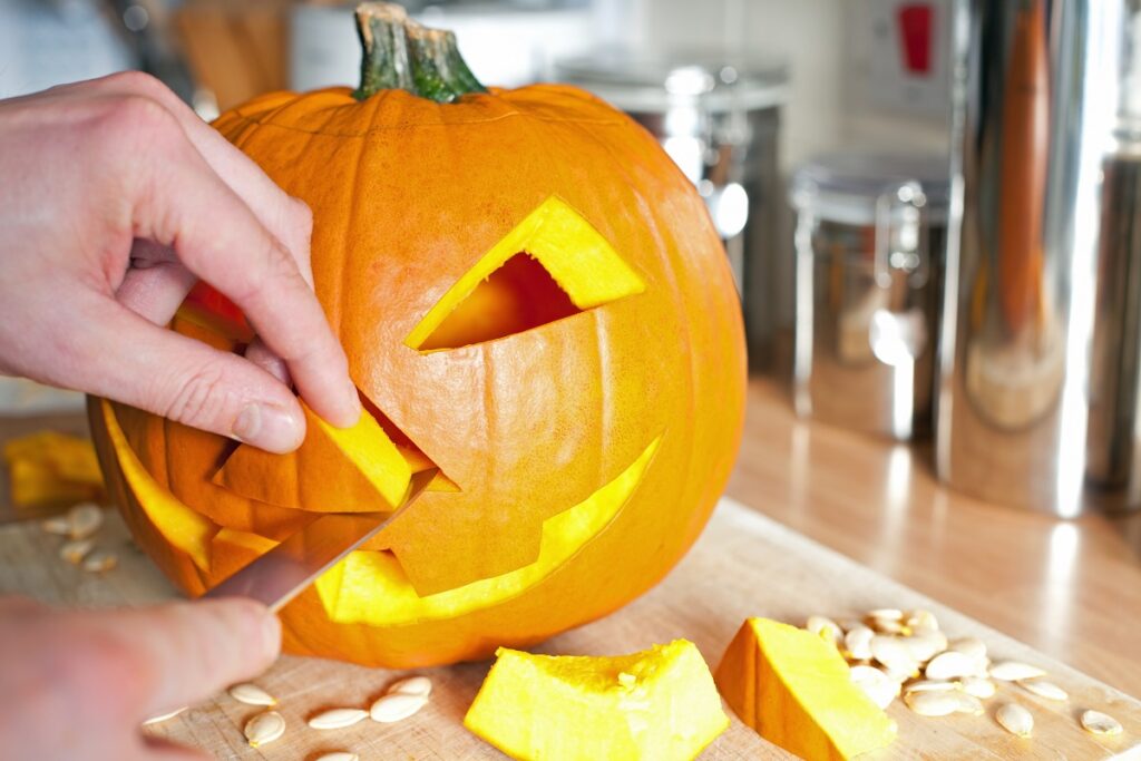 A close-up of hands carving a classic jack-o'-lantern from a pumpkin during a team building event. The partially carved pumpkin features a smiling face, with cut-out pieces and pumpkin seeds scattered on the table, highlighting the creative and hands-on nature of the activity.