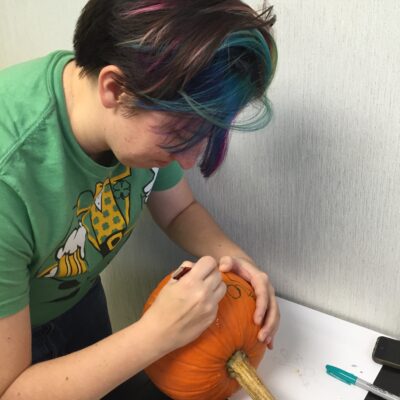 A person concentrates on drawing a design onto a pumpkin during a team building pumpkin carving event. They carefully sketch the outline, preparing the pumpkin for carving, showcasing creativity and attention to detail in the activity.