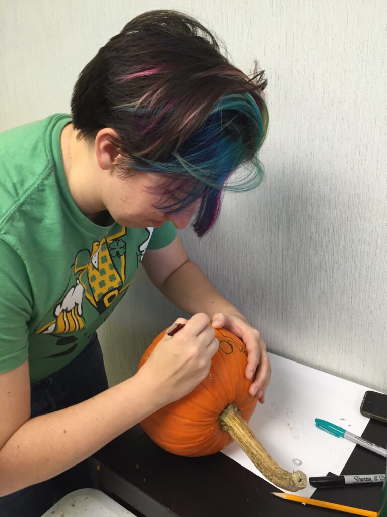 A person concentrates on drawing a design onto a pumpkin during a team building pumpkin carving event. They carefully sketch the outline, preparing the pumpkin for carving, showcasing creativity and attention to detail in the activity.