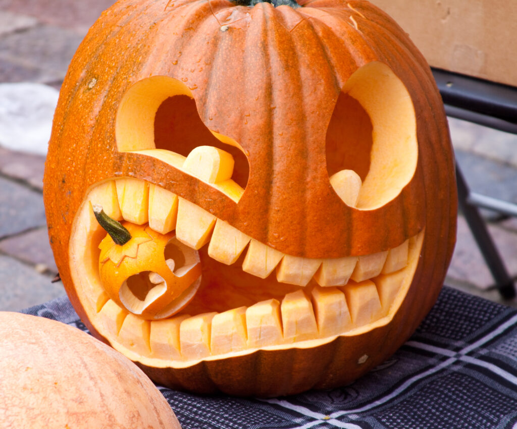 A creatively carved jack-o'-lantern featuring a unique design of a large pumpkin with exaggerated teeth, 'biting' a smaller carved pumpkin. The playful and intricate carving showcases the artistic and humorous approach to pumpkin carving.