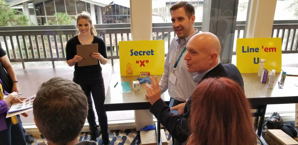 Participants at a Foodbank Showdown team building event engage in grocery-themed game challenges at different stations labeled 'Secret X' and 'Line 'em Up' to win items for donation to families in need.