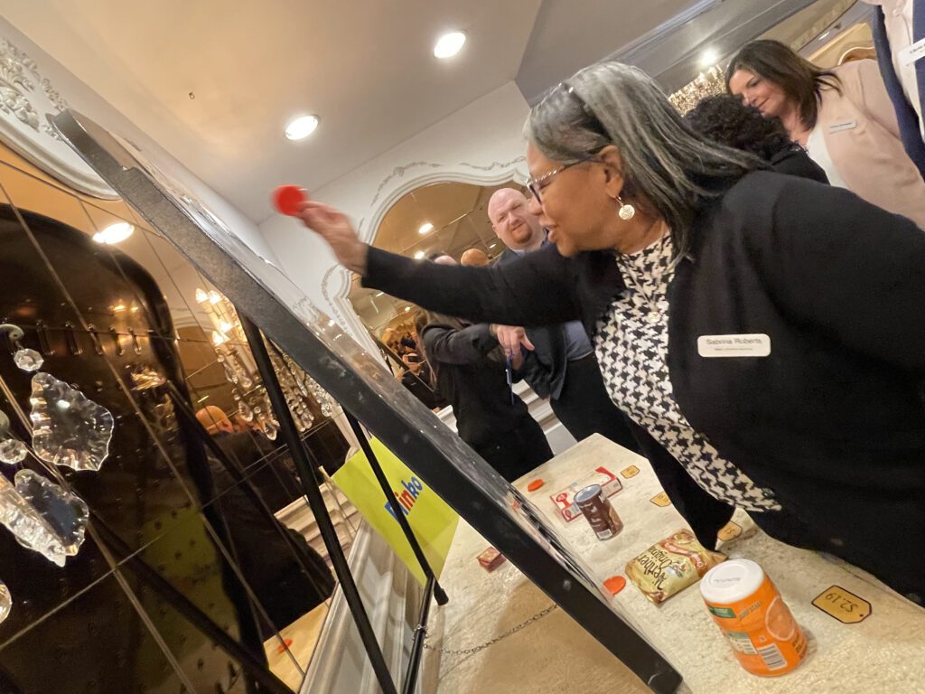A participant enthusiastically throws a red disc onto a game board during the Foodbank Showdown team building event. In the background, grocery items are visible, showcasing the event's purpose of gathering goods for charity.