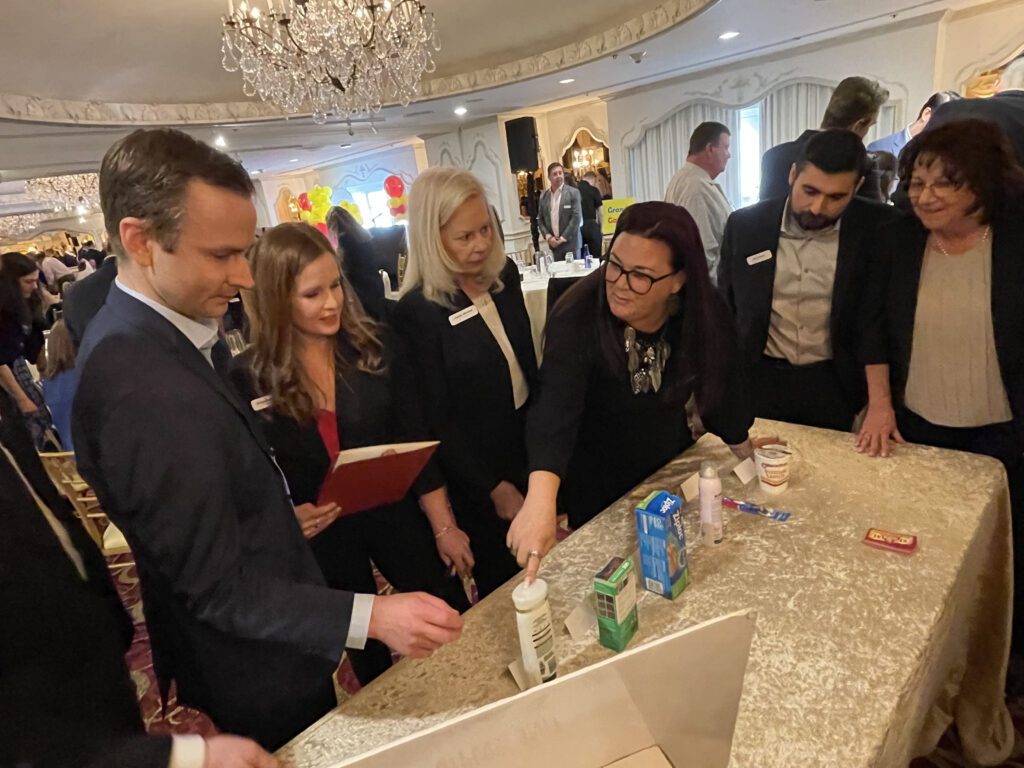 A team of participants collaborates during the Foodbank Showdown team building event, working together to select and purchase grocery items for charity. The elegant ballroom setting adds to the engaging atmosphere as they strategize for success.