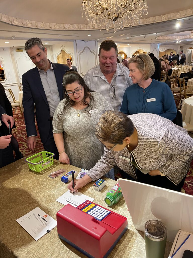 Participants gather around a table engaging in a fun grocery-themed challenge during the Foodbank Showdown team building event. A red toy cash register and mini grocery items are on the table, emphasizing the game's interactive and charitable aspect.