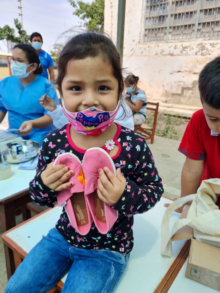 A young girl wearing a colorful mask smiles as she holds a pair of pink TOMS shoes that she received through the Paint Soles for Souls charitable team building event. In the background, healthcare workers assist other children.