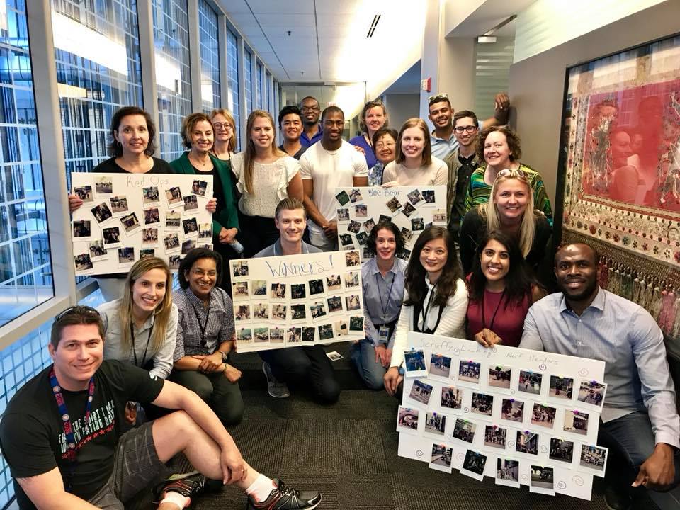 A large group of team members posing together in a hallway, proudly displaying their collage boards filled with Polaroid photos from a scavenger hunt team building event. The boards are labeled with team names and showcase the creative challenges completed during the activity. The group smiles in celebration, in a fun and energetic atmosphere.