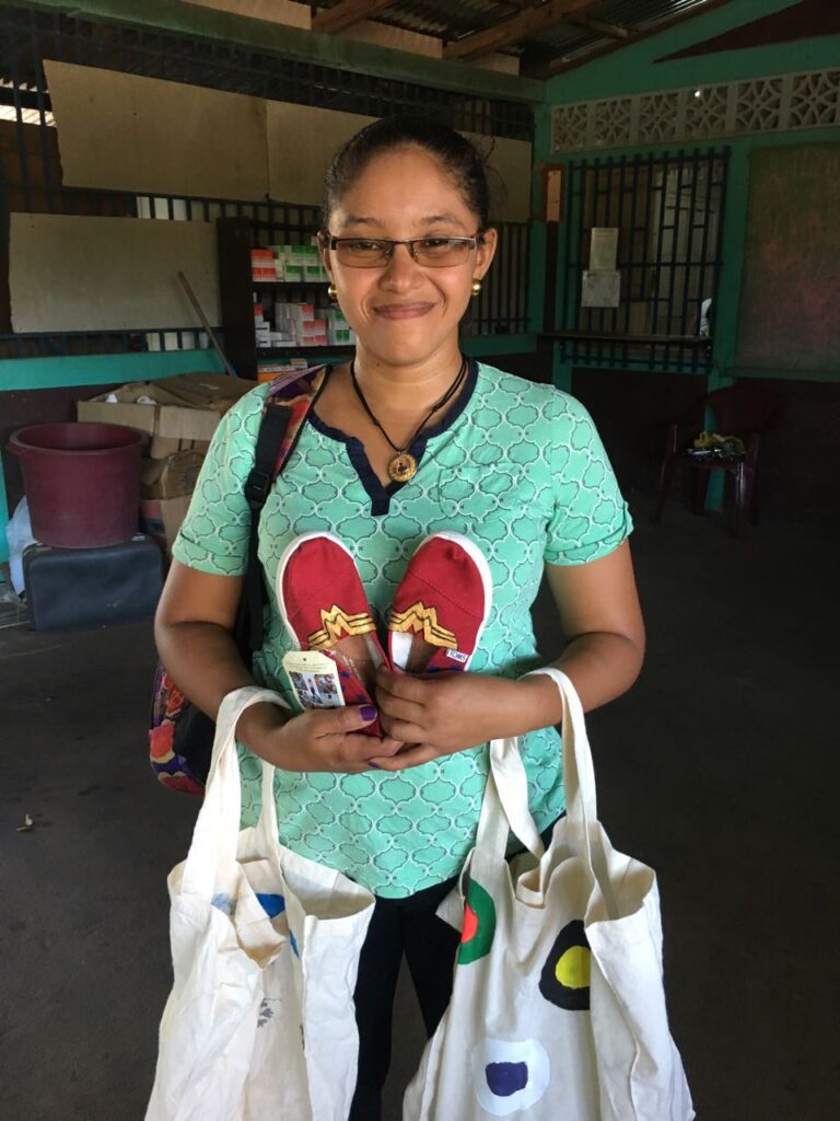 A smiling woman holds a pair of red, hand-painted TOMS shoes featuring a Wonder Woman design, along with decorated tote bags, which were donated through the Paint Soles for Souls charitable team building event.