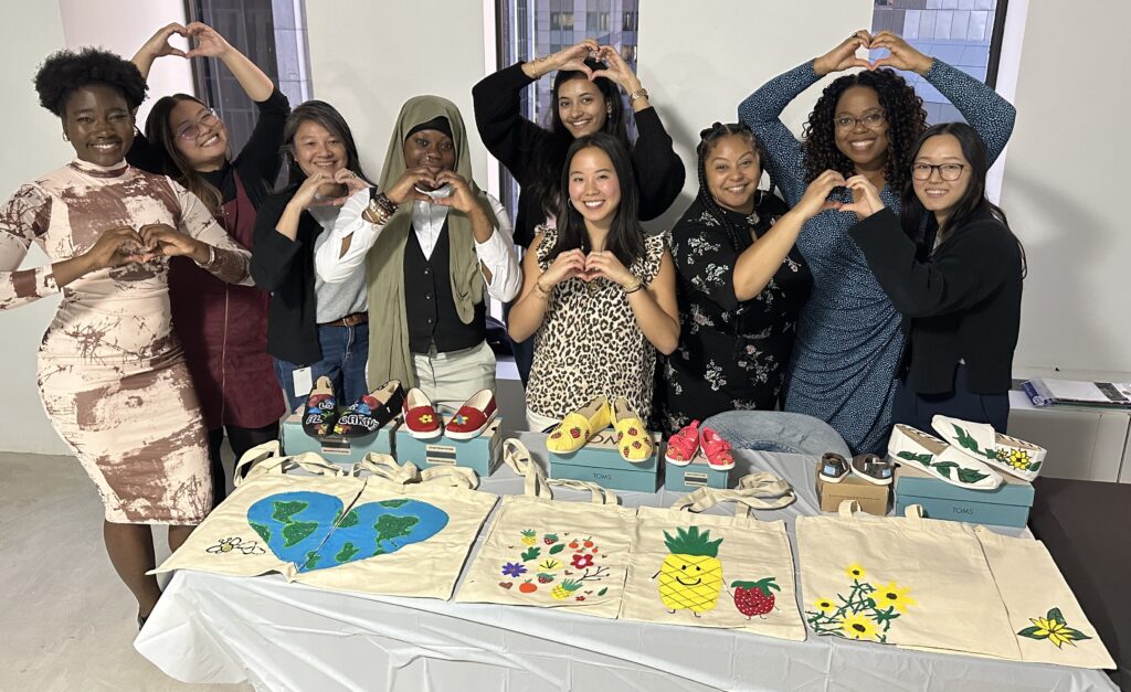 A group of smiling participants stands and makes heart gestures with their hands. They are displaying hand-painted TOMS shoes and tote bags created during the Paint Soles for Souls charitable team building event.