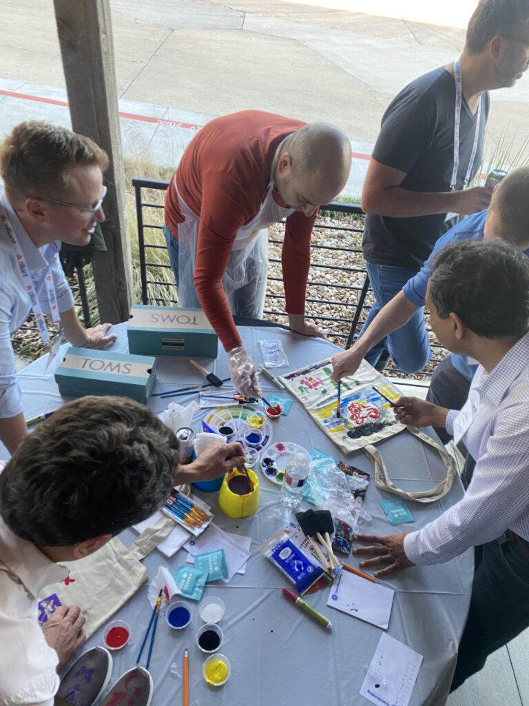 A group of participants collaborates on painting TOMS shoes and tote bags during the Paint Soles for Souls charitable team building event. The table is filled with art supplies, including paints, brushes, and TOMS shoeboxes, as participants creatively design items that will be donated.