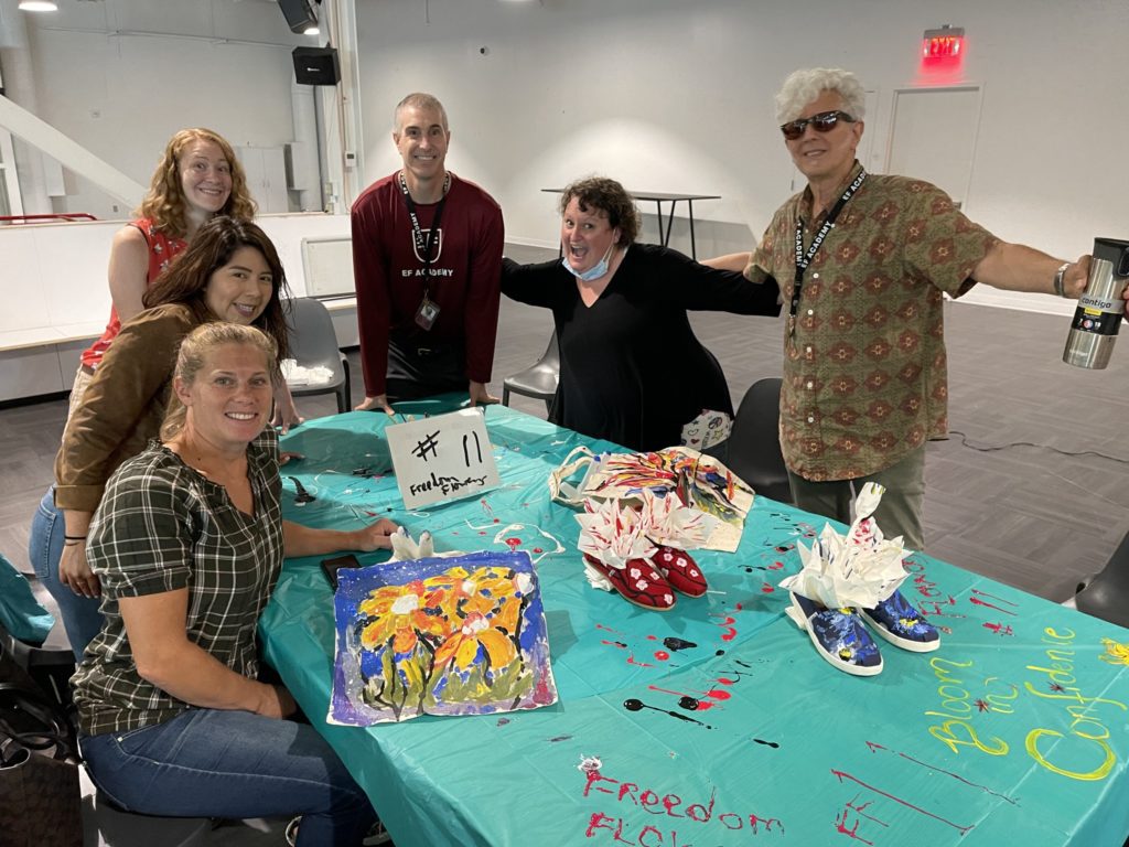 A group of participants poses around a table displaying their hand-painted TOMS shoes and decorated tote bags during the Paint Soles for Souls charitable team building event. The colorful items, featuring floral and abstract designs, will be donated to children in need in South America. The event encourages creativity, teamwork, and giving back to the community.