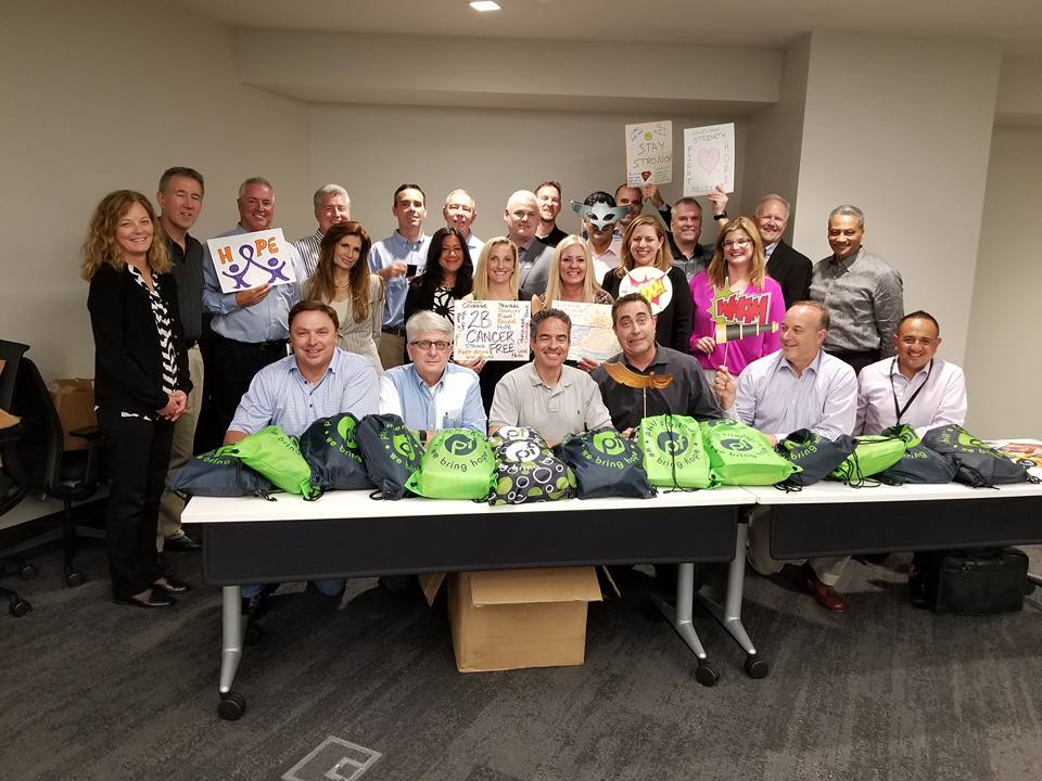 A group of people posing in a conference room behind a table filled with green drawstring bags. Some individuals are holding up supportive posters with words like 'Hope,' 'Stay Strong,' and 'You Are Cancer-Free.' The group is smiling, showing support for cancer patients as part of a team building event focused on creating care packages.
