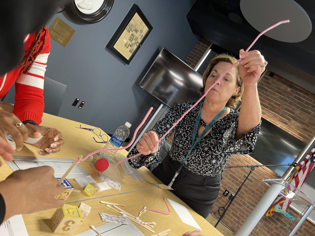A woman carefully balancing a long pink pipe cleaner while participating in a hands-on team building challenge. She is surrounded by small wooden blocks, sticks, and other materials on a table.
