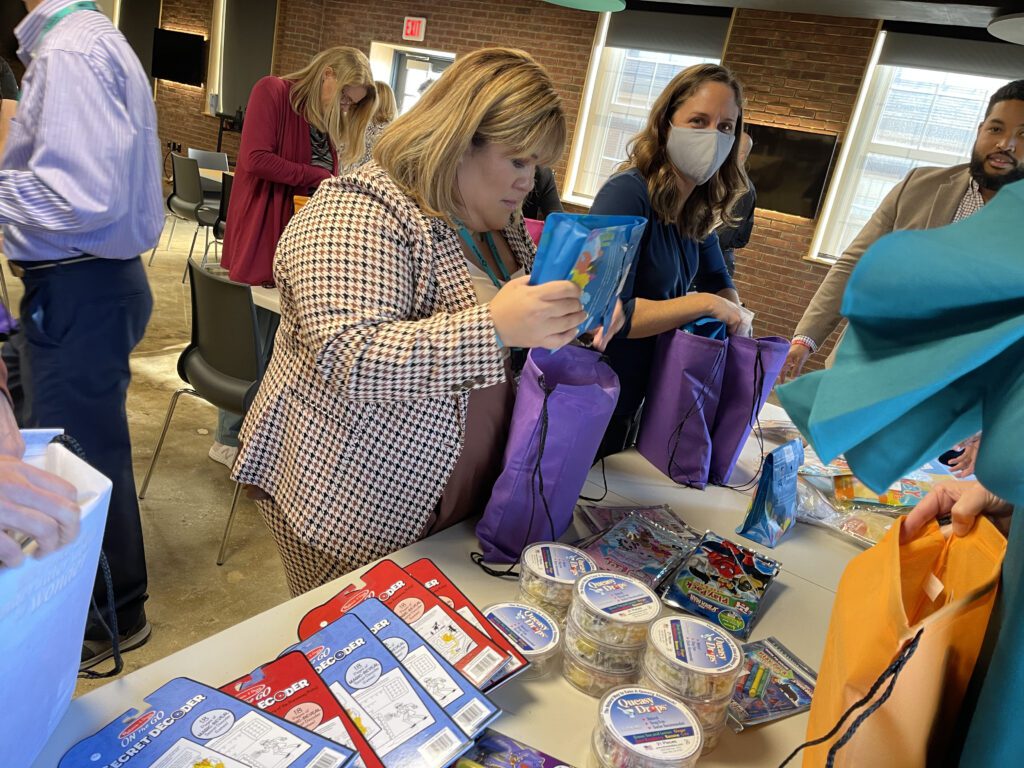 A group of people are participating in a team building activity where they are assembling care packages. A woman in the foreground is holding a purple bag and carefully placing an item inside. Various supplies are laid out on the table to put together care-kits to give to cancer patients