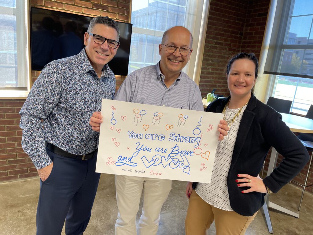 Three people standing together and smiling while holding a large, hand-drawn poster. The poster reads: 'You are Strong, You are Brave and Loved.' The poster is decorated with heart and balloons.