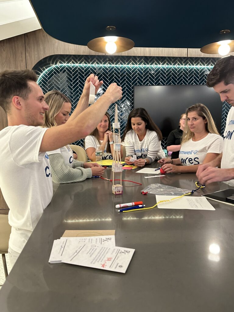 A group of people engage in a team building activity around a table. They are working together to build a structure using various materials such as string and sticks, with a focused and collaborative atmosphere.