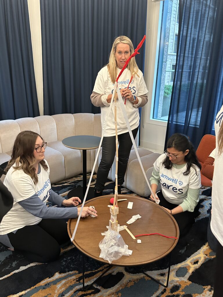 Three team members are participating in a team building challenge. They are gathered around a small round table, constructing a tall tower using wooden sticks, tape, and string. Two participants are sitting on the floor working on the base, while one stands, adjusting the top of the structure.